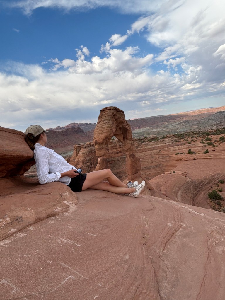 A woman in a white long-sleeve shirt and shorts sits on a rock ledge, overlooking a red rock landscape with a prominent rock arch under a cloudy sky.