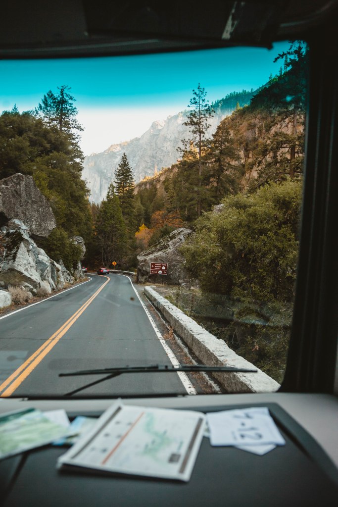 View of a winding road through a forested area, seen from inside a vehicle, with roadside maps visible on the dashboard.