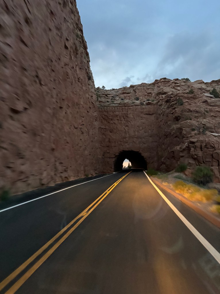 A road leading through a rocky tunnel with a light at the end, surrounded by rugged terrain under a cloudy sky.