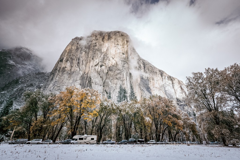 A towering granite cliff surrounded by autumn trees and snow, with parked vehicles along a road in the foreground.