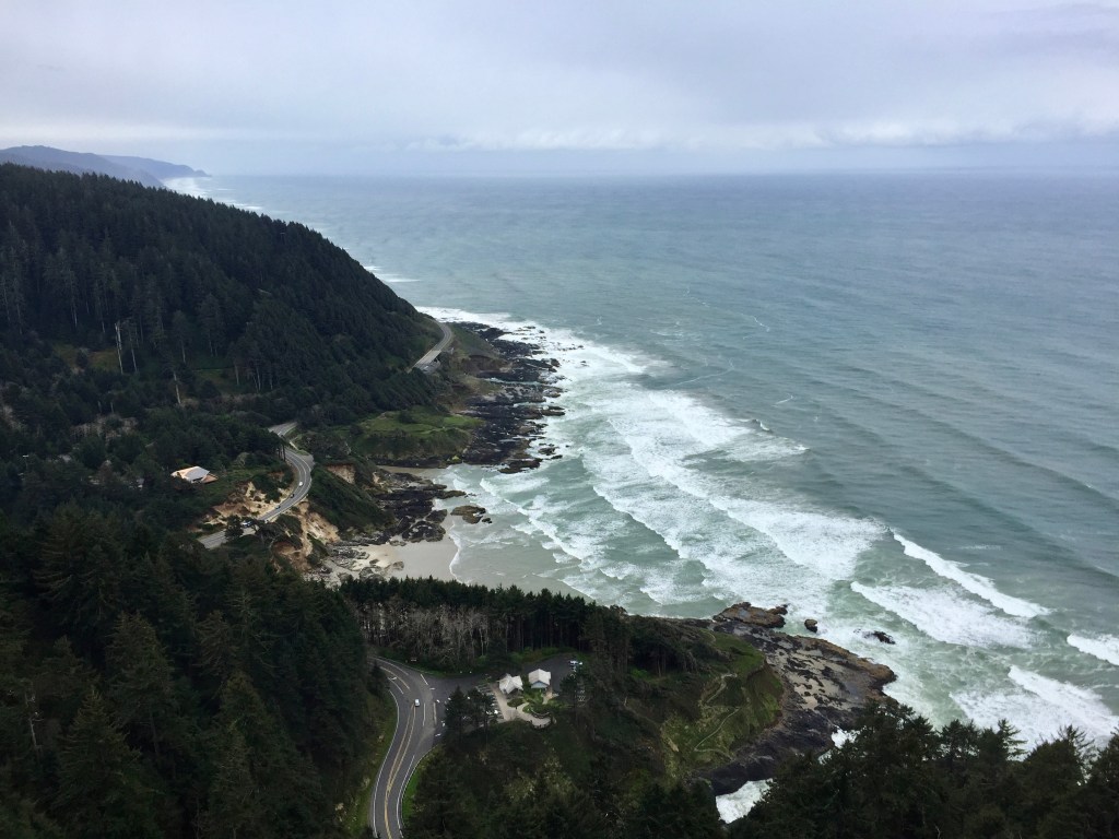 A panoramic view of a rugged coastline with waves crashing on rocks, surrounded by lush green forests and a winding road along the shore.